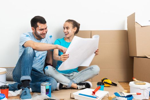 Happy couple sitting on floor with construction blueprint. Home remodeling and house interior redesign. Construction tools and materials lying on floor. Young family studies renovation project of flat