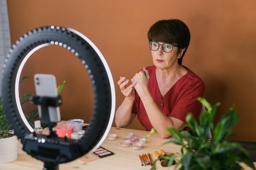 Middle-aged woman talking on cosmetics with makeup eye shadows and blush palette and brushes while recording her video. Mature female making video for her blog on cosmetics