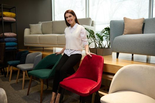 Cute sales assistant in casual clothes, standing near velour stylish chairs in the furniture shopping mall, smiles looking at camera while presenting the new collection of upholstered furniture