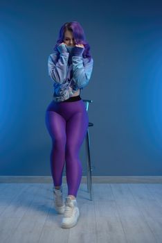 a girl in stylish purple sportswear and with purple hair poses sexually on a bar stool