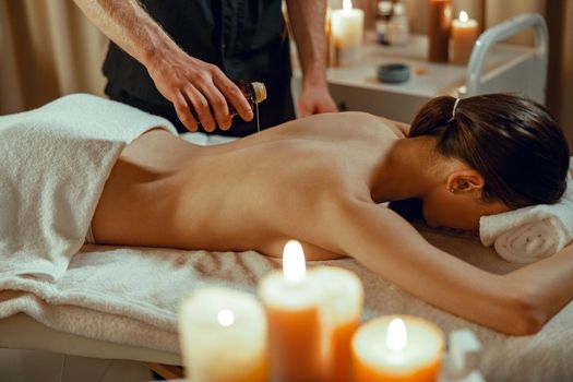 Masseur man therapist pouring warm herb infused oil on female back