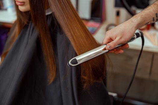 Closeup of hairdresser hand holding hair straightener and making hairstyle for female client
