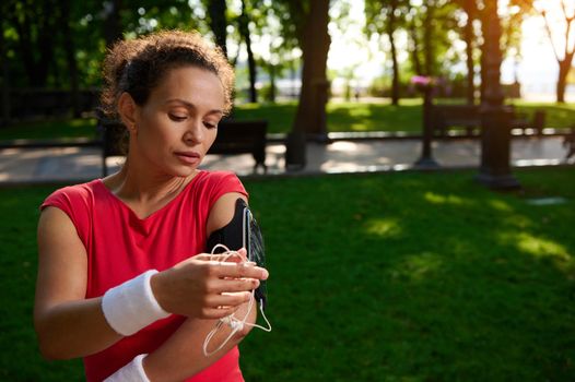 Young athletic woman, a runner in a bright red t-shirt, connects headphones to a mobile phone and sets up the application before jogging in the park on a sunny summer day