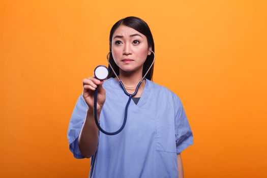 Concerned asian nurse using stethoscope to consult sick patient while on orange background.