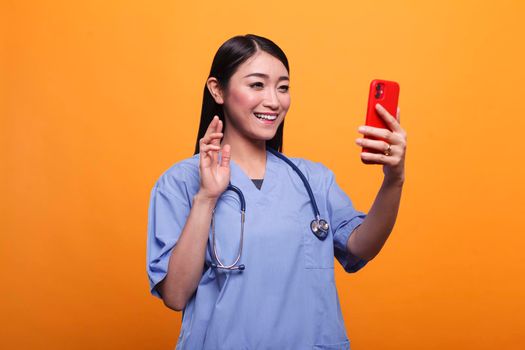 Healthcare clinic nurse wearing medical instrument waving at teleconference videochat call. Caregiver in remote telehealth videocall with patient while having online consultation on orange background.