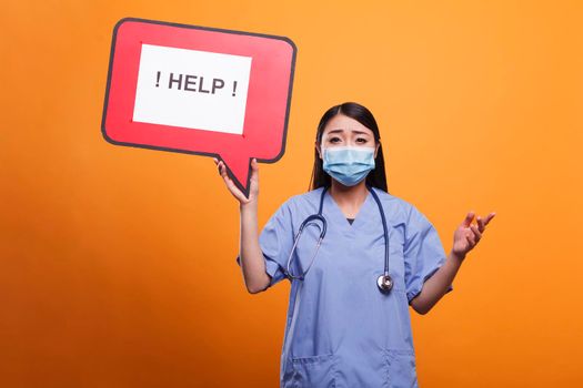 Healthcare clinic nurse holding cardboard speech bubble sign while wearing protective mask and medical uniform. Scared nurse wearing stethoscope and medical clothing asking for help.