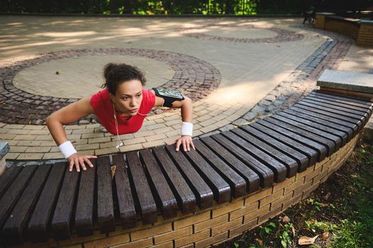 Determined active woman exercising outdoor, pushing up from wooden bench, performing full body cross training outdoor