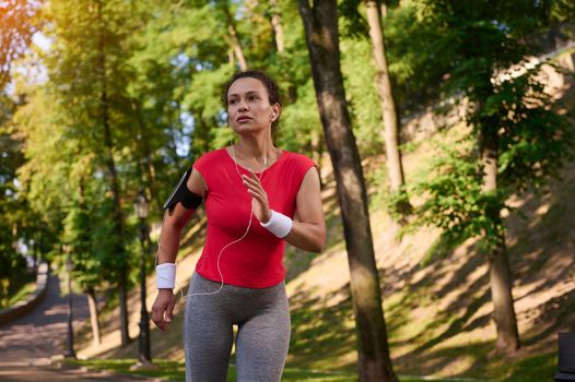 Attractive strength muscular multi ethnic woman in tight sportswear with earphones and smartphone holder running on the footpath in the city park