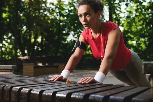 Determined Hispanic female athlete doing pushups from the wooden bench in the city park during functional cross training in the morning