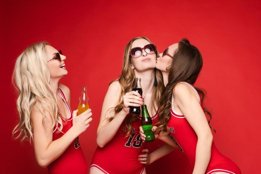 Beautiful and happy female friends in bright red swimsuits with chilled drinks in their hands on a red background, studio.