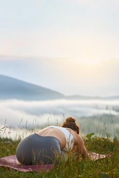 Back view of young slim woman practicing yoga in hills. Flexible female sitting on legs, bending on yoga mat in mountains. Concept of new age religion.
