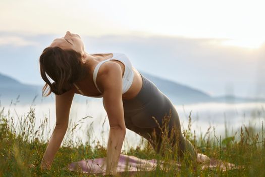 Side panoramic view of young woman practicing yoga pose in hills. Female standing in plank with closed eyes wearing sportswear outdoors. Concept of harmony and health.