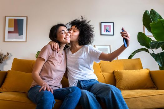 Young lesbian couple taking selfie at home using mobile phone. Girlfriend gives kiss on the cheek.