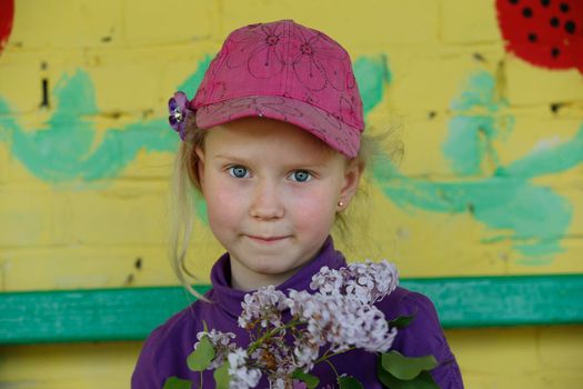 . A beautiful little girl in a hat holds a twig of lilacs and smiles.