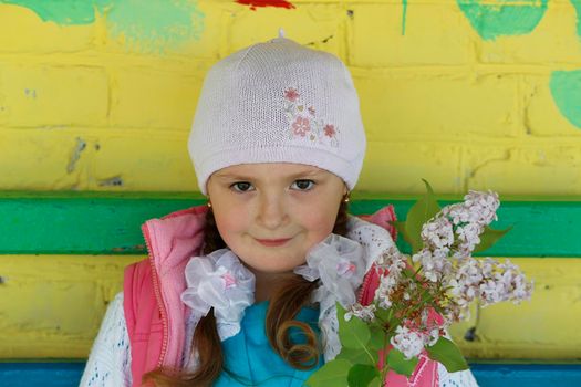 A beautiful little girl in a knitted hat holds a sprig of lilacs and smiles.