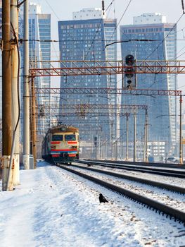 An electric train moves on rails against the backdrop of a cityscape of skyscrapers in a winter haze.