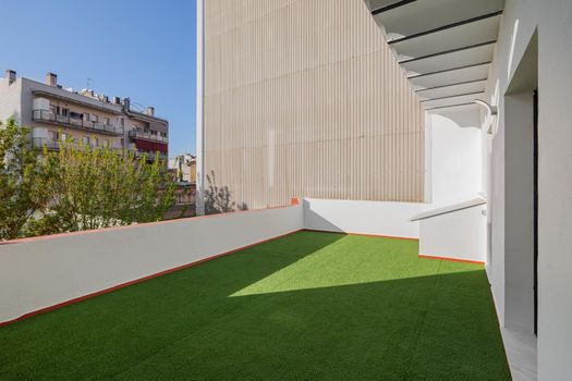 Empty sunny terrace of refurbished apartment with artificial green grass lawn. View to the street and blue sky in the center of Barcelona