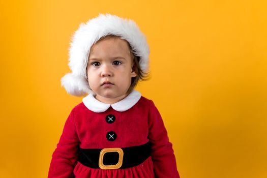 Serious Portraite Cute Happy Cheerful Chubby Baby Girl in Santa Hat Looking On Camera At Yellow Background. Child Play Christmas Scene Celebrating Birthday. Kid Have Fun Spend New Year Time Copy Space