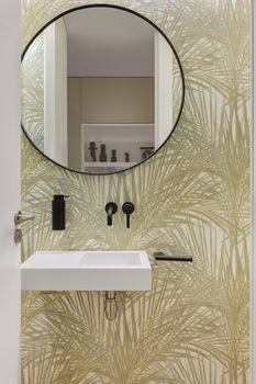 Bathroom with wallpaper with plant leaves texture, small wash basin and round mirror. Minimalist interior.