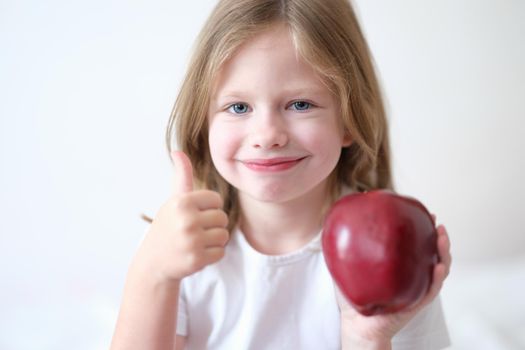 Little girl holding red apple in hands and showing thumbs up gesture