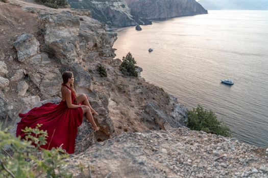 A girl with loose hair in a red dress sits on a rock rock above the sea. In the background, the sea. The concept of travel.