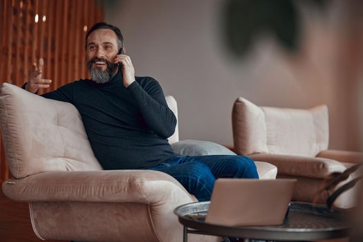 Smiling bearded man sitting in armchair and chatting on phone