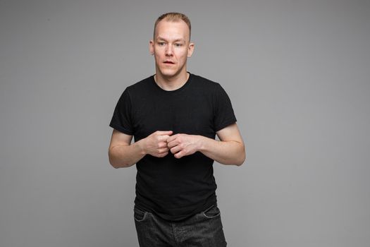 attractive man with short fair hair wearing a black t-shirt and jeans keeps hands together and talking
