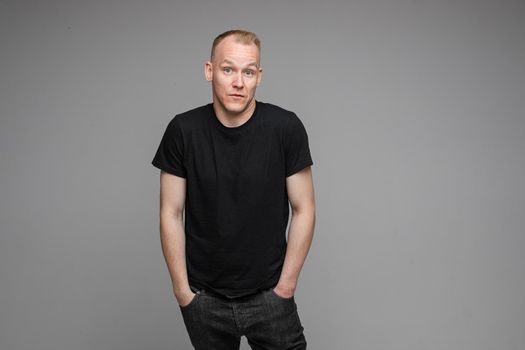 attractive man with short fair hair wearing a black t-shirt and jeans keeping hands in pockets and surprising of something