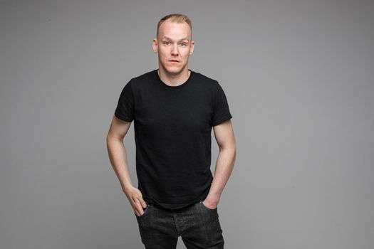 attractive man with short fair hair wearing a black t-shirt and jeans keeping hands in pockets and surprising of something