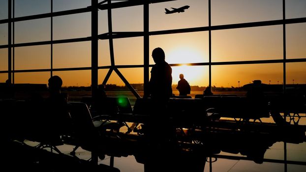 Silhouette of a tourist guy watching the take-off of the plane standing at the airport window at sunset in the evening. Travel concept, people in the airport.