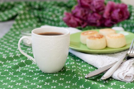 Black coffee in a white sophisticated cup, delicious dietary cheesecakes from home-made farmer cheese for breakfast, a bouquet of purple tulips on a green tablecloth.