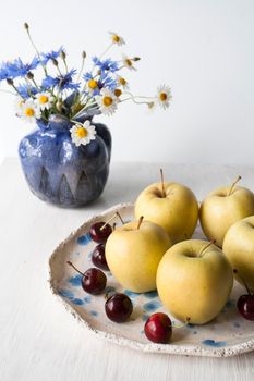 Yellow apples and juicy cherries lie on a plate of clay. Nearby is a pigeon vase with field flowers. Composition on a white wooden background.