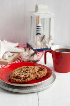 Oatmeal cookies with cranberries lie on a red plate, next to a cup of black coffee, a flashlight, seashells and an old notebook. The composition is on a striped tablecloth and white wooden background.