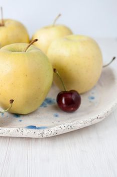 One juicy red cherry and a few yellow tasty apples in a clay plate on a white wooden background. The view is close.
