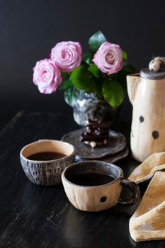 Two cups of black coffee, black chocolate, a yellow teapot and a vase with pink roses on a dark background.