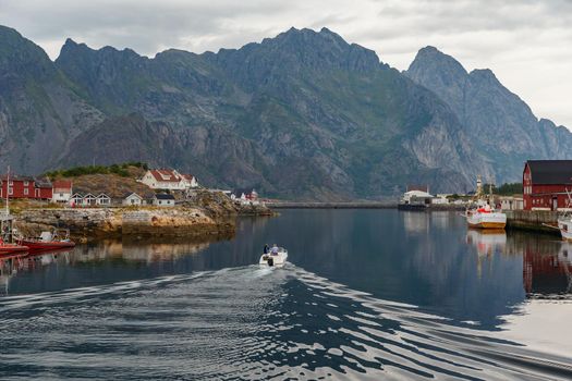Norwegian seascape, cityscape of the town Henningsvaer, a small boat moves between peninsulas, sail boat, rocky coast with dramatic skies, classic view of Norwegian houses on the slopes