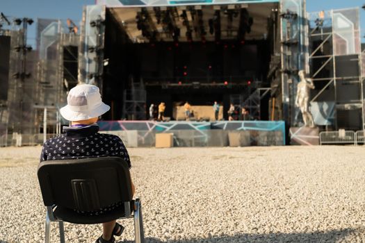 in front of the huge open stage where the art festival will take place on a huge empty platform in front of the stage, the director of the show is sitting on a chair and watching the rehearsal going on on stage