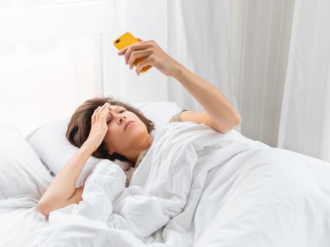 Sleepy woman looks on smartphone screen. Checking e-mail box right after waking up. Using wireless technology in bed.