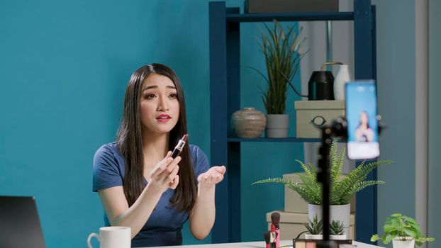 Asian woman recommending beauty product on vlogging camera, filming makeup cosmetics review with broadcast equipment. Influencer recording lifestyle vlog with lipstick, online video.