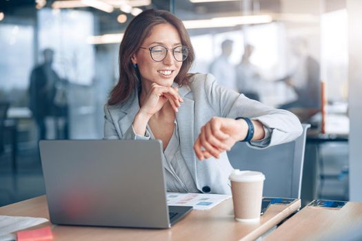 Positive businesswoman wearing eyeglasses looks at her wristwatch at modern office