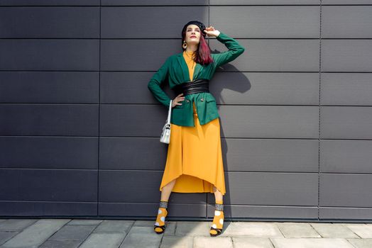 Young woman in bright clothes, yellow skirt and green jacket. Yellow socks in sandals, beret on the head, hair with the color of magenta. Caucasian female fashion model standing against gray wall background, open with copy space.