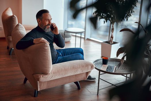 Happy bearded man sitting near window and chatting on phone