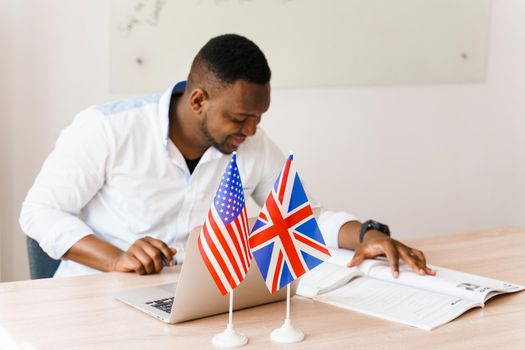 English and British flags in front. Black handsome translator uses his laptop for online work according social distancing