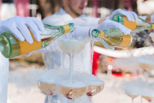 Waiter pours champagne from 2 bottles into crystal glasses with dry ice and white smoke close up. Sparkling wine pouring in wine glass