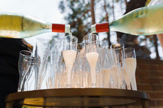 2 waiter pours champagne in disposable plastic wine cup. Pouring sparkling wine into wine glass on welcome drink zone in the forest. Catering for business people.