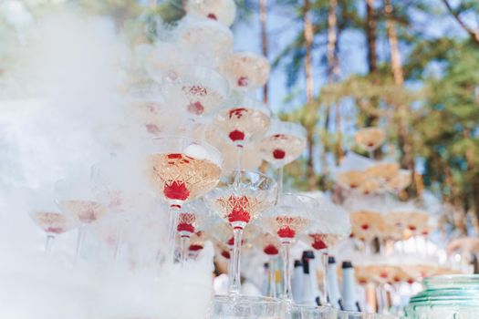 Pyramid of champagne with smoke of dry ice and sparkling wine and red cherry inside. Catering for wedding ceremony and business people.