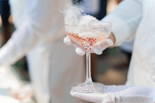 Waiter in white gloves holds wine glass with sparkling wine, red cherry, and white smoke of dry ice and gives to customer