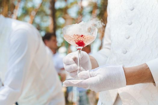 Waiter in white gloves holds wine glass with sparkling wine, red cherry, and white smoke of dry ice and gives to customer