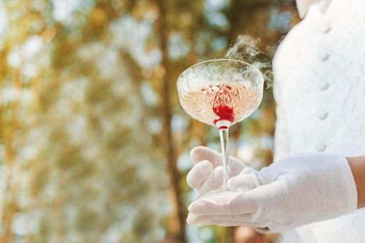 Waiter in white gloves gives wine glass with shampagne, red cherry, and white smoke of dry ice and gives to customer. Catering for wedding ceremony.