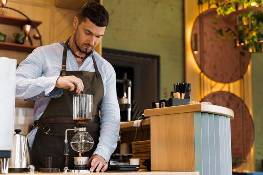 Handsome bearded barista mix freshly ground coffee in syphon device for coffee brewing in cafe. Syphon alternative method of making coffee.